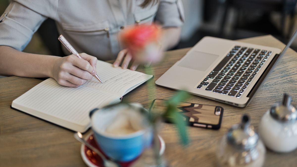 A notebook, pen and laptop on a desk beside a cup of coffee.